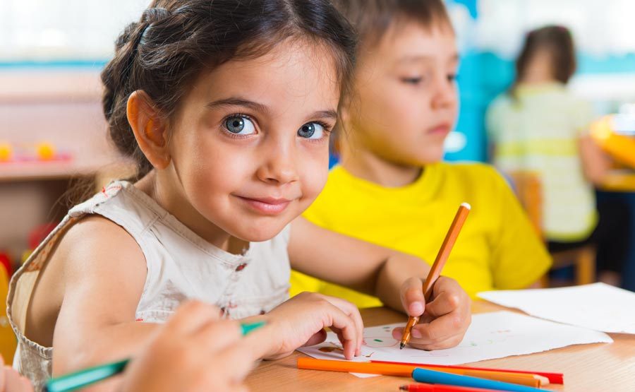 Young child with big eyes smiling, looking at camera while drawing at a desk in a childcare centre setting